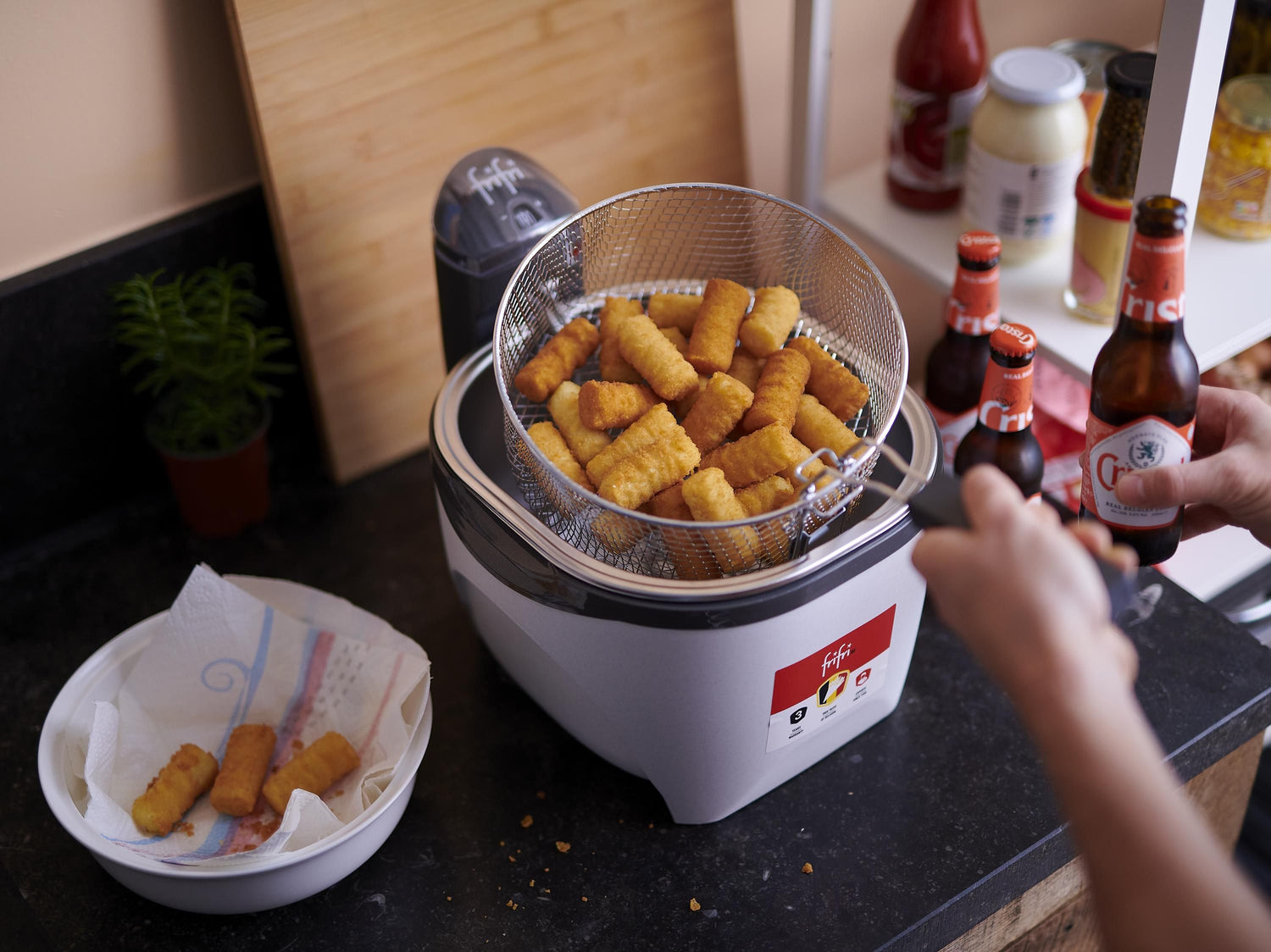 Une personne fait frire des snacks panés dans une friteuse sur un comptoir de cuisine, avec des bouteilles de bière et un bol contenant des snacks frits à proximité.