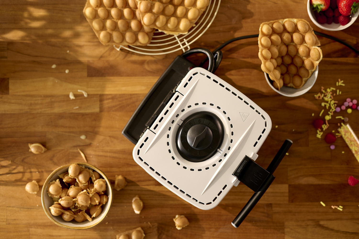 Vue de haut des plaques Gaufrier Violet + 8 sur une table en bois, entourées de gaufres à bulles, d'un bol de morceaux de gaufres, de baies et de paillettes colorées.