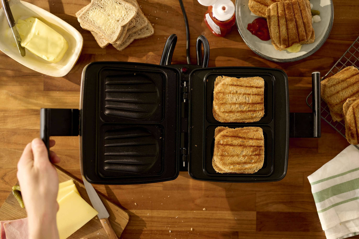Une personne tient le manche d'un Gaufrier Gris + 8 plaques avec deux sandwichs grillés à l'intérieur ; du beurre, du pain, du fromage et des sandwichs cuits reposent sur le comptoir en bois.