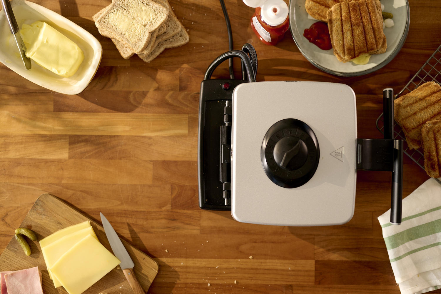Vue de dessus d'une table en bois avec un appareil à croque-monsieur, du beurre, du pain et des ingrédients pour la garniture.