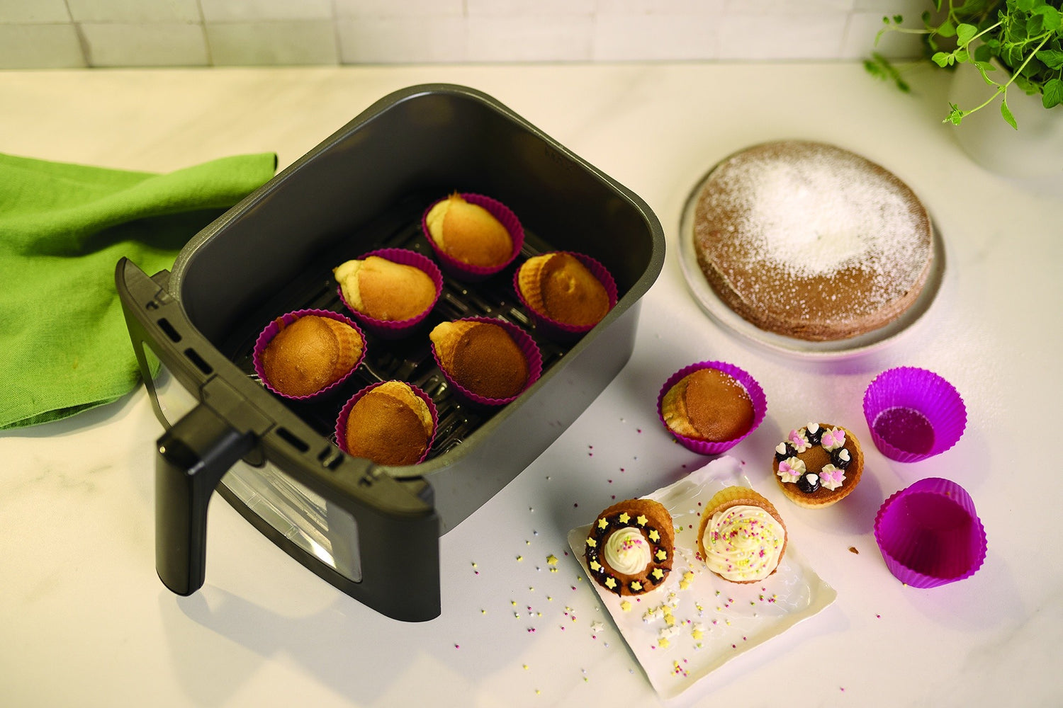 Vue rapprochée d'une friteuse à air avec des cupcakes en silicone violet, un gâteau saupoudré de sucre glace, et une serviette verte sur une table blanche.