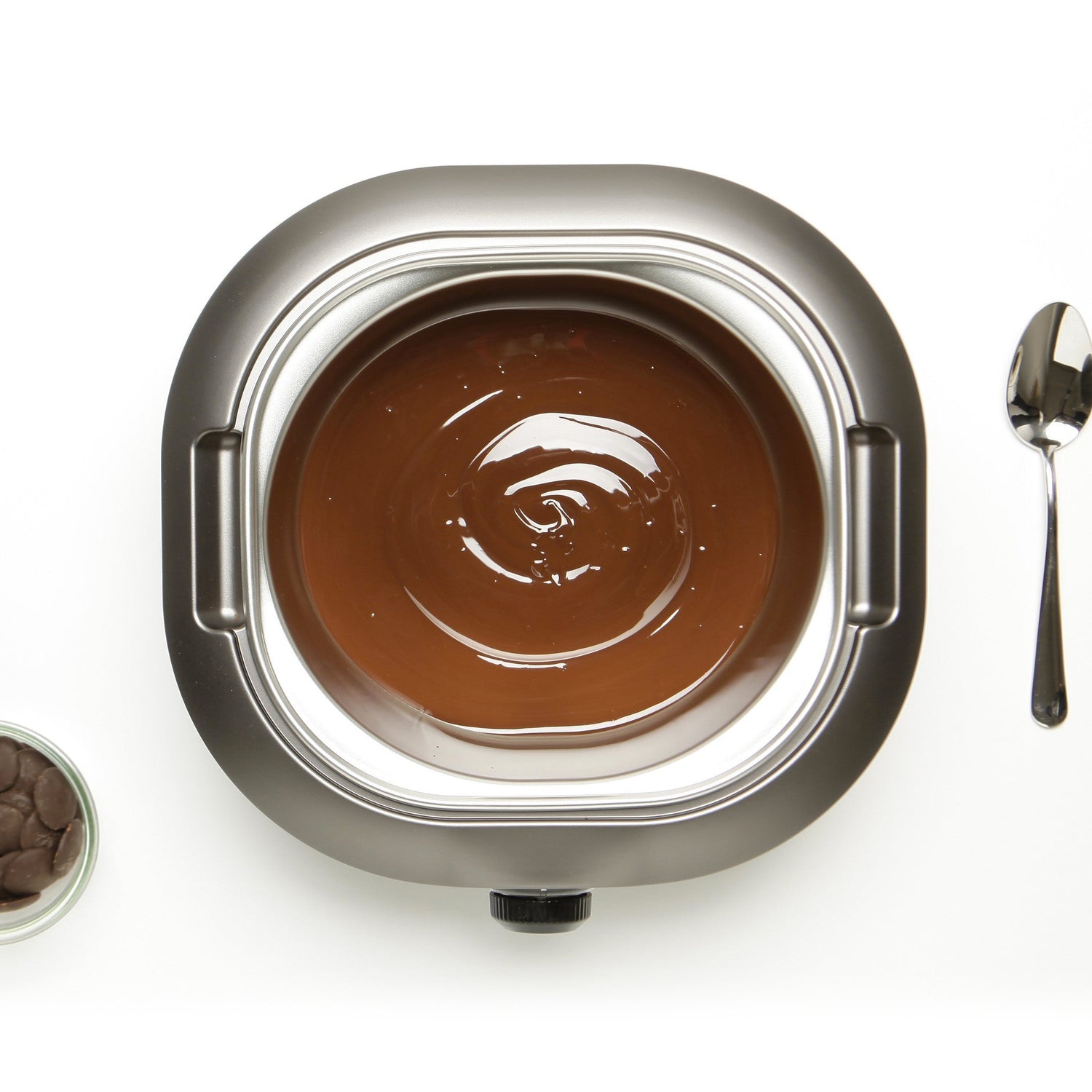 Vue aérienne du chocolat fondu dans un fondoir électrique La Chocolatière en acier inoxydable, avec une cuillère et un petit bol de pépites de chocolat à proximité.