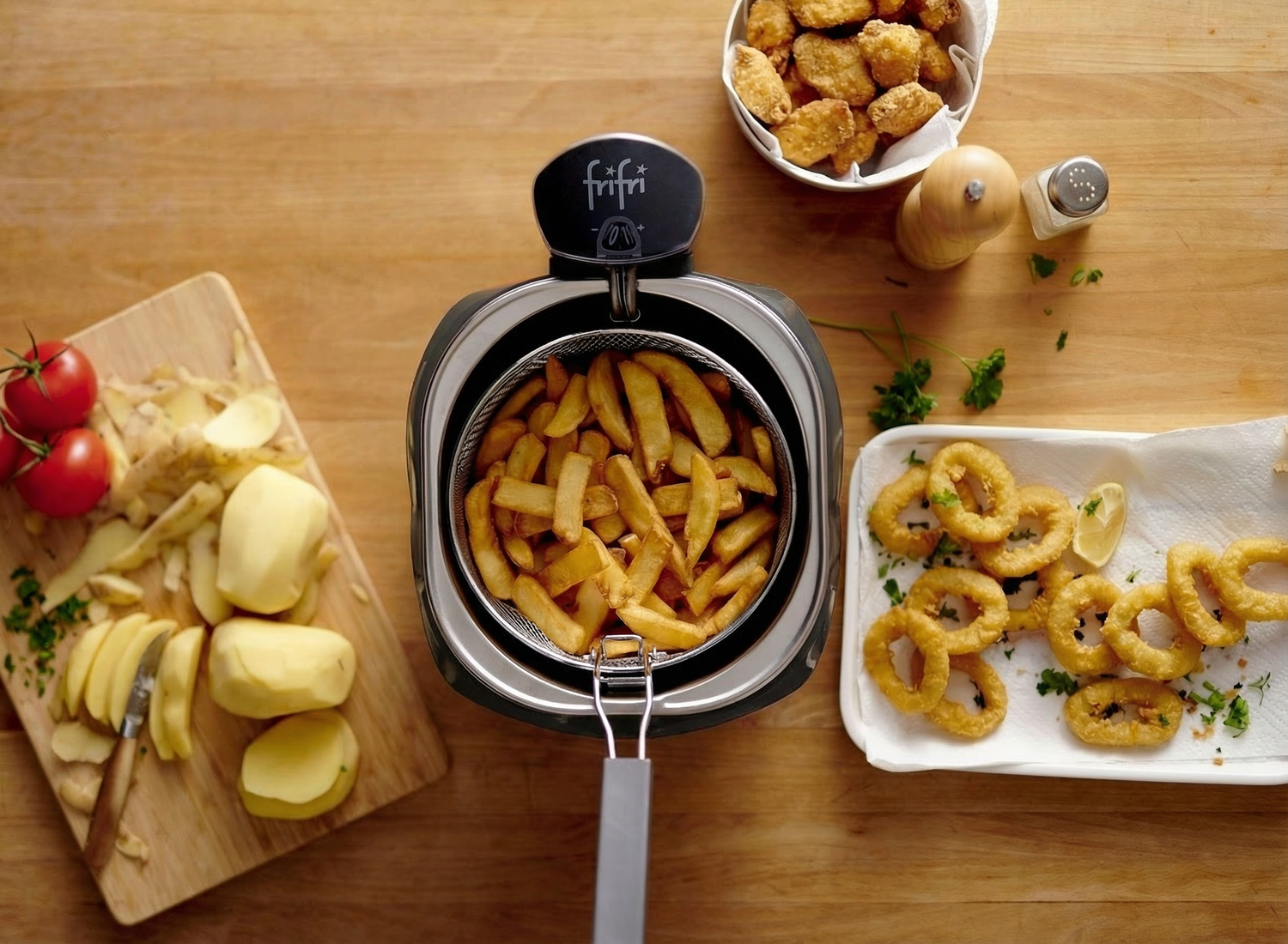 Une friteuse contenant des frites cuites est posée sur une table en bois, entourée de pommes de terre crues, de tomates, de nuggets frits et de rondelles d'oignon panées sur une serviette en papier.