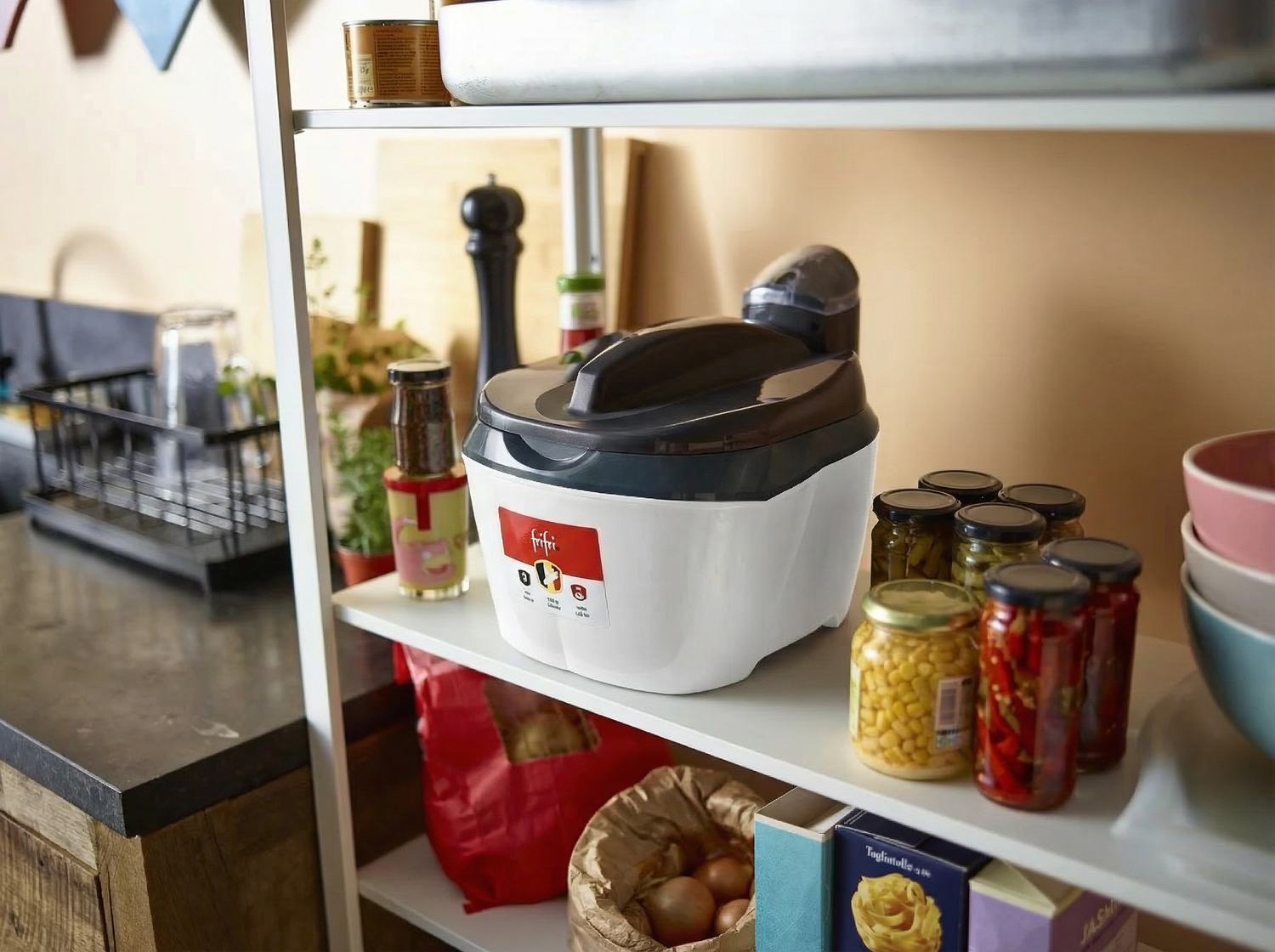 Un cuiseur de riz électrique blanc trône sur une étagère de la cuisine, entouré de bocaux, de conserves, de bols et d'autres accessoires de cuisine.