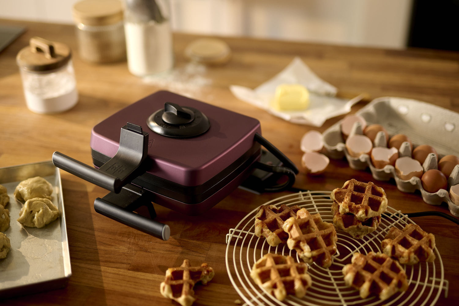 Un gaufrier est posé sur un comptoir en bois, entouré d'œufs, de beurre, de farine, de pâte à biscuits et de gaufres fraîchement préparées sur une grille de refroidissement.