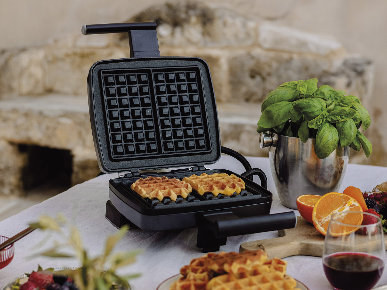 Un gaufrier ouvert avec deux gaufres à l'intérieur est posé sur une table, entouré de fruits frais, d'un verre de vin rouge et d'un plant de basilic en pot.