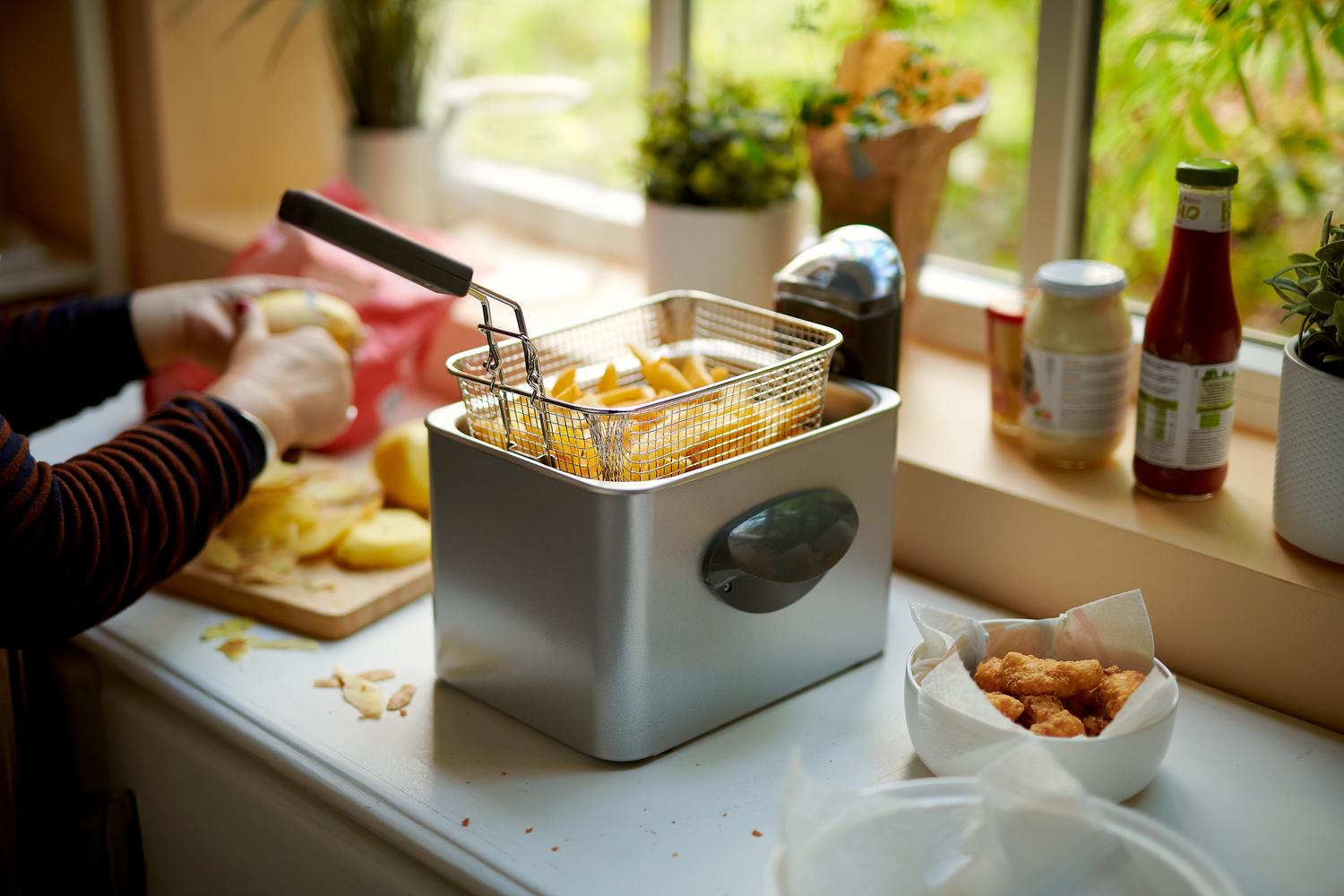 Une personne prépare des pommes de terre à côté d'une friteuse avec des frites coupées à l'intérieur ; des aliments cuits et des condiments sont posés sur le comptoir voisin, près d'une fenêtre avec des plantes en pot.