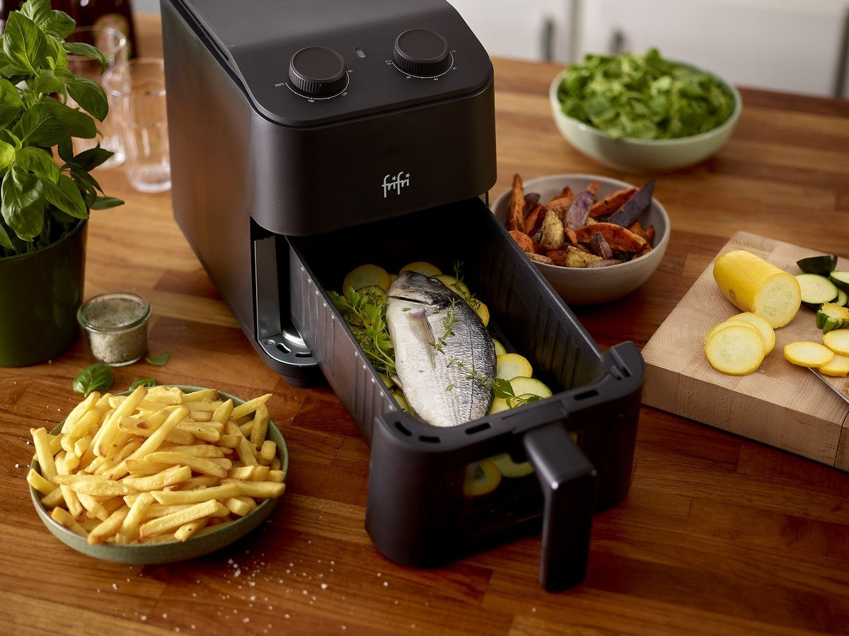 Une friteuse avec un poisson entier et des légumes en tranches dans le panier trône sur le comptoir d'une cuisine, entourée de bols de frites, de frites de patates douces et de salade.