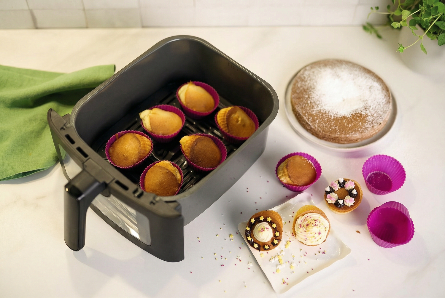 Le panier de l'Airfryer Duo Slim contient plusieurs cupcakes dans des moules roses, un gâteau rond au sucre en poudre, des cupcakes décorés et une serviette verte sur un plan de travail blanc.