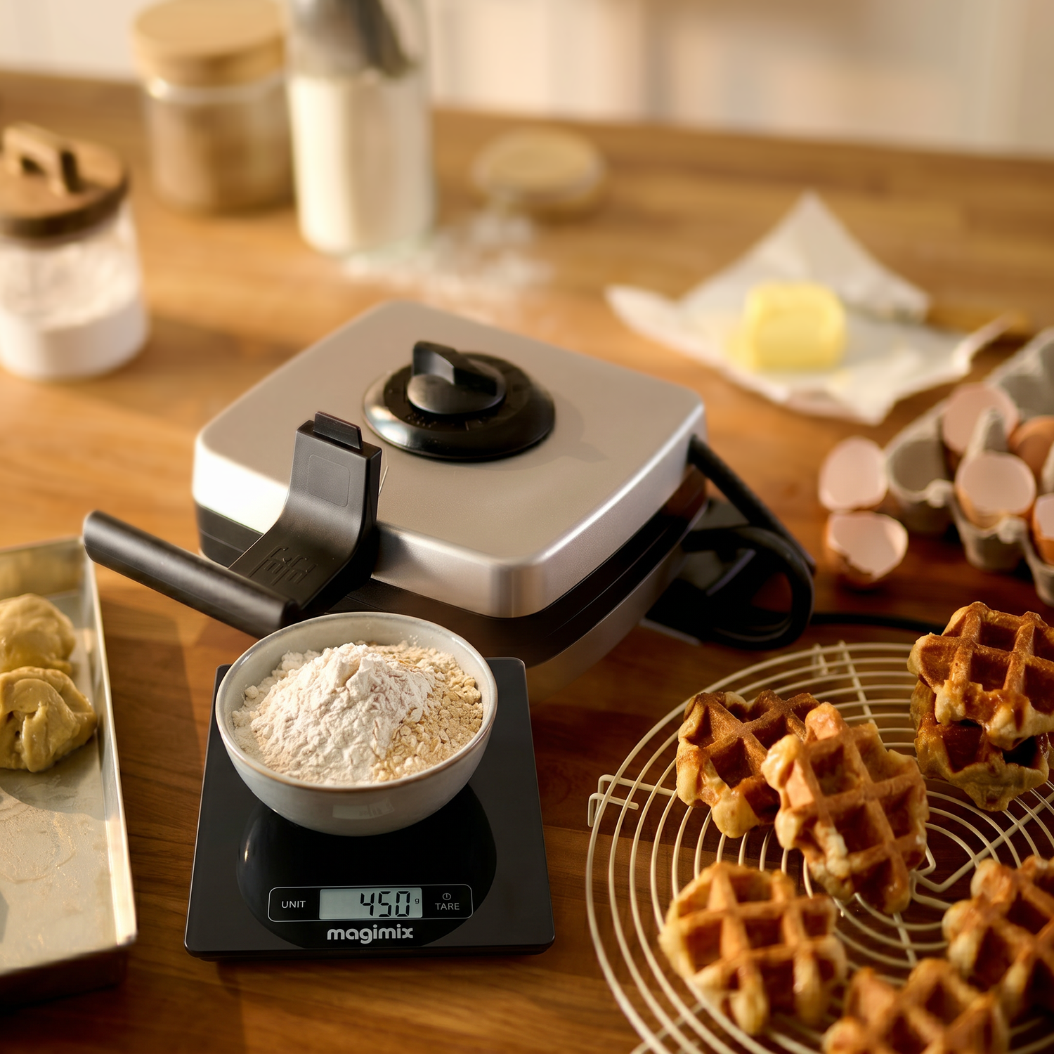 Un plan de travail avec un gaufrier Magimix, Balance de précision contenant de la farine, un bac à pâte, des coquilles d'œufs, du beurre et des gaufres fraîches prêtes pour des recettes sucrées et salées.