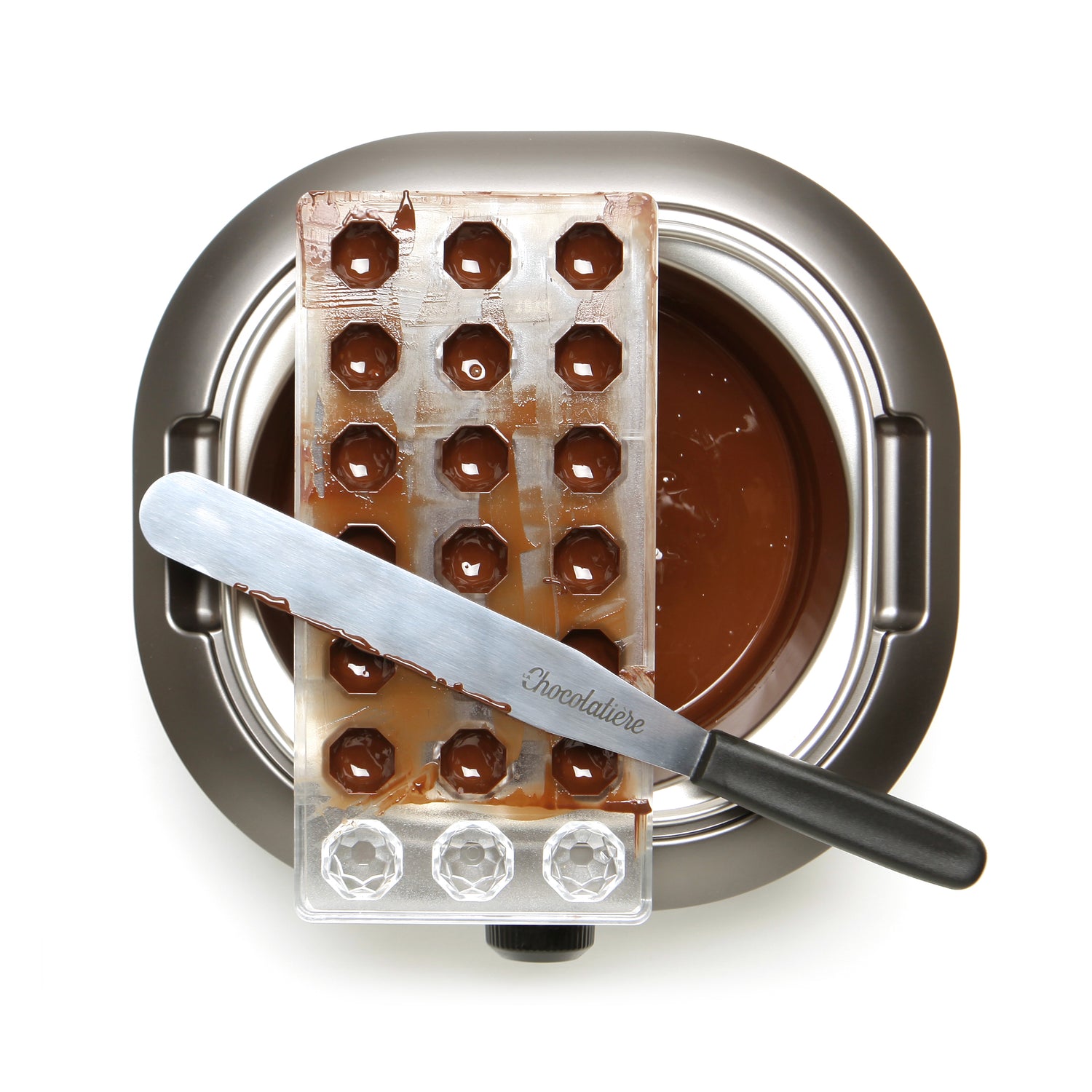 La Chocolatière + Kit de Pâques comprend un moule à pralines en métal, des chocolats et une spatule posée sur un bol de chocolat fondu - parfait pour préparer de délicieuses friandises de Pâques maison.