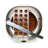 La Chocolatière + Kit de Pâques comprend un moule à pralines en métal, des chocolats et une spatule posée sur un bol de chocolat fondu - parfait pour préparer de délicieuses friandises de Pâques maison.