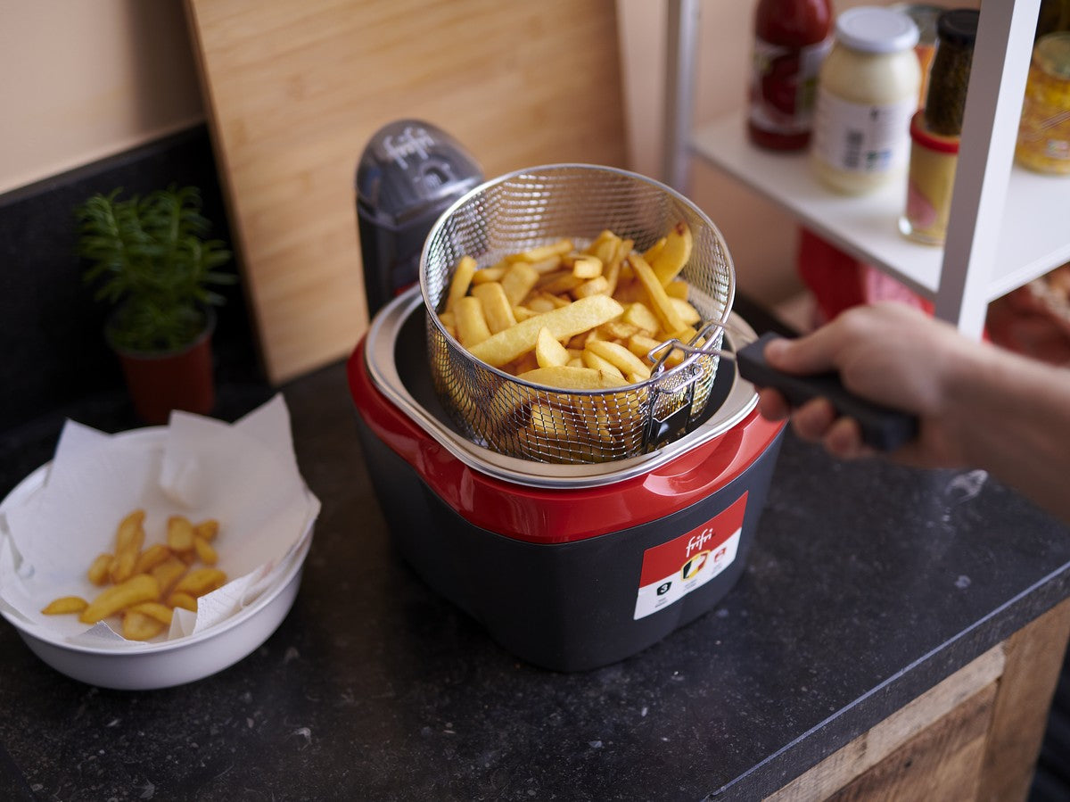 Une personne sort un panier de frites cuites d'une friteuse rouge et noire posée sur un comptoir de cuisine, avec un bol de frites et des condiments à proximité.