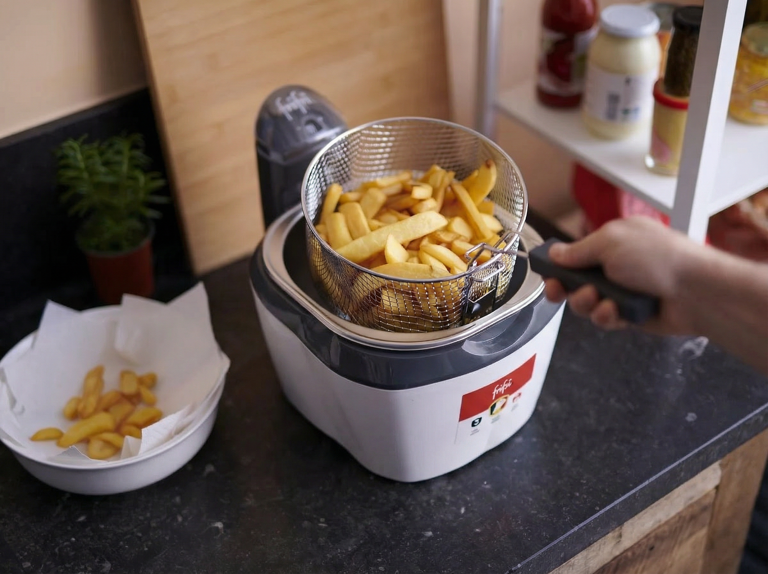 Une personne utilise une friteuse pour cuire des frites dans une cuisine, avec un bol de frites cuites et des condiments visibles sur le comptoir à proximité.